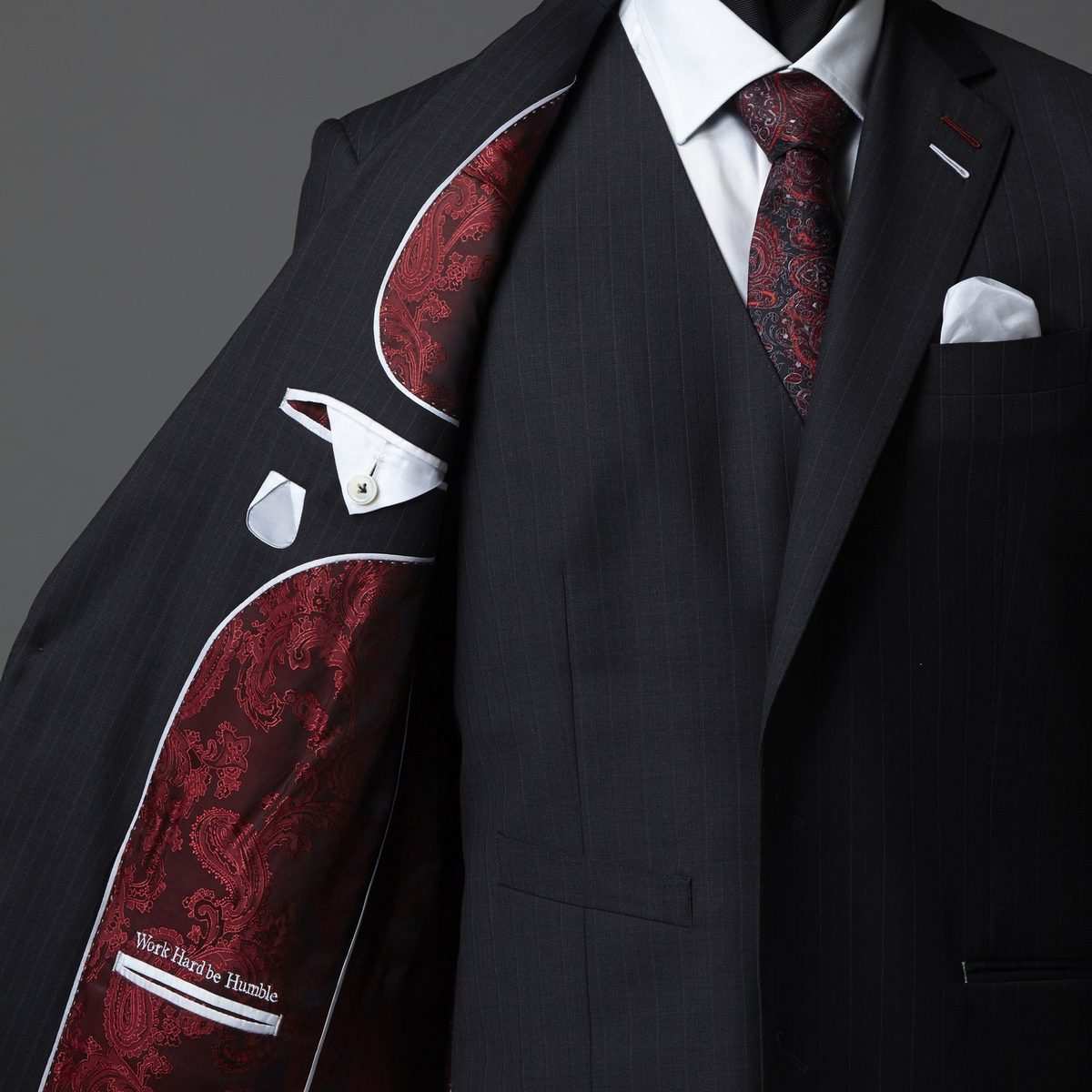 Intérieur d'une veste anthracite : doublure jacquard paisley bordeaux bordée d'un passepoil blanc, inscription « Work Hard be Humble » brodée au-dessus de la poche intérieure