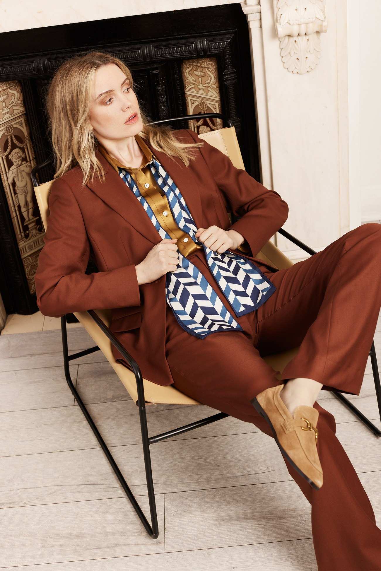 Tailleur pantalon rouille et foulard de soie à chevrons, assise dans un fauteuil de cuir