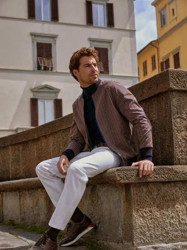 Blouson bordeaux et pantalon clair, assis sur un muret de pierre