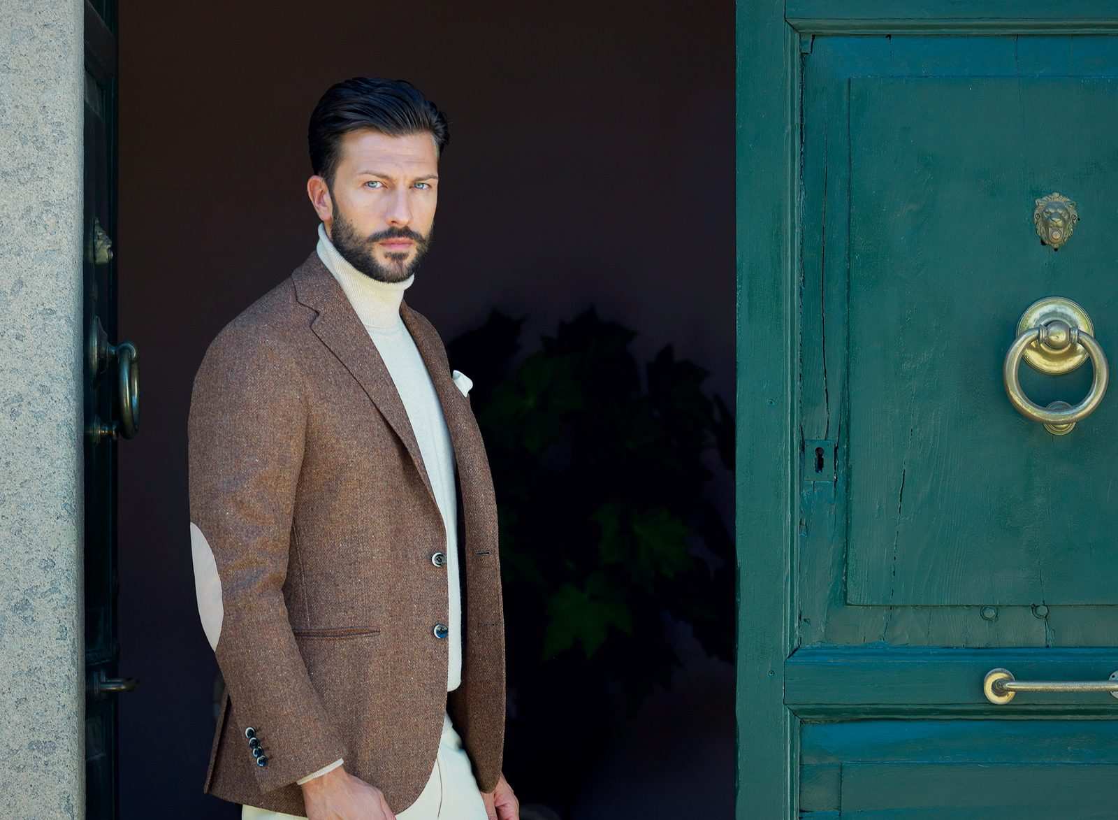 Homme en veste de tweed brune à coudières et col roulé écru, devant une porte verte
