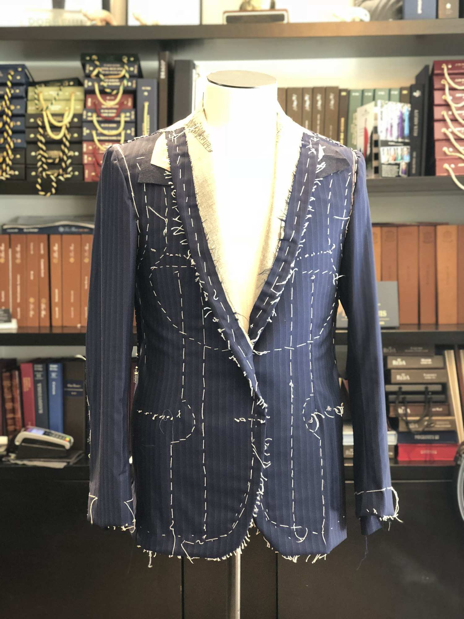 Veste bleu marine à rayures montée en bâti d'essayage, faufils blancs apparents, sur un buste devant les liasses du salon